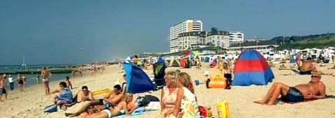 Auf einem hellen Sandstrand liegen viele Leute und sonnen sich. Manche haben Strandmuscheln aufgebaut. Im Hintergrund ist ein sehr hohes Gebäude zu sehen. (Foto: SWR - Print aus der Sendung)