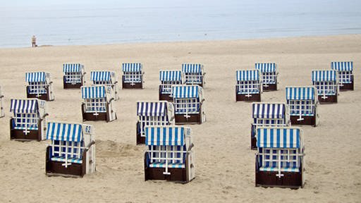 Auf einem Sandstrand stehen viele blau-weiß gestreifte Strandkörbe, im Hintergrund ist das Meer mit einem einsamen Spaziergänger zu sehen. (Foto: Huber, www.pixelio.de)
