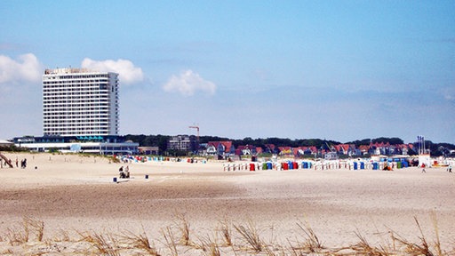 Auf einem Sandstrand stehen viele bunte Strandkörbe, dahinter sind rechts kleine Häuser zu sehen, links im Bild ein Hochhaus mit dem Schriftzug „Hotel Neptun“.