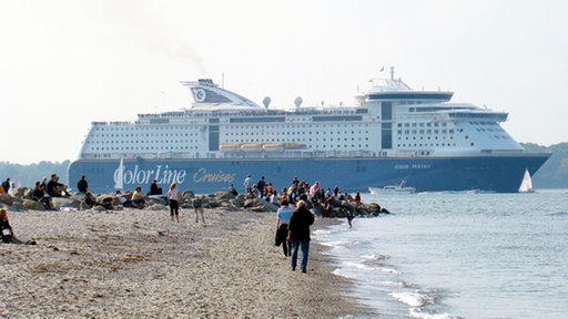 Ein sehr großes Fährschiff mit blauem Rumpf und weißem Aufbau läuft ins Meer aus. Im Vordergrund Strand mit vielen Schaulustigen.