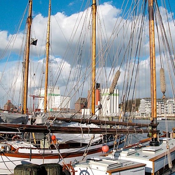 Mehrere Segelschiffe liegen nebeneinander im Wasser, im Hintergrund sind hohe Häuser zu sehen.