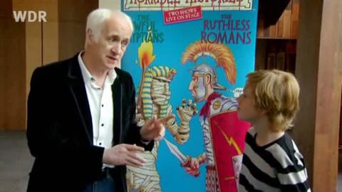 Terry Deary steht mit einem Jungen vor einem Plakat mit dem Buchcover der "Horrible Histories"