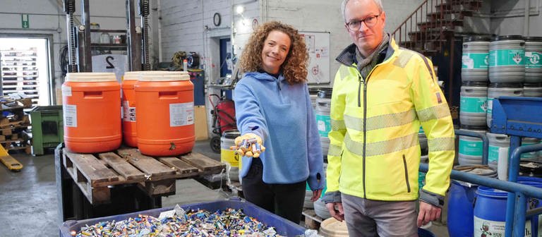 neuneinhalb-Reporterin Gesa und Florian Clever stehen vor einer großen blauen Plastikwanne voller Altbatterien in einer Recycling-Anlage. Im Hintergrund stehen bunte Plastikfässer voller Batterien. Gesa hält eine Handvoll Batterien in Richtung Kamera. (Foto: WDR, tvision)