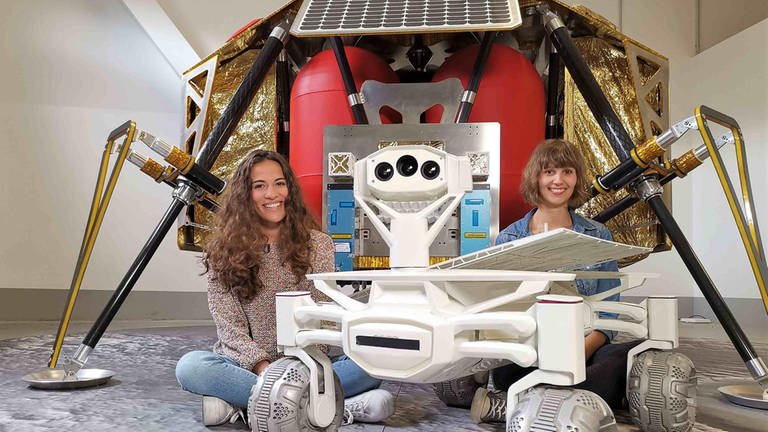 neuneinhalb-Reporterin Mona und Wissenschaftlerin Nadine sitzen vor einer Raumsonde, neben ihnen steht ein Rover, der eines Tages auf dem Mond umherfahren soll. (Foto: WDR, tvision)