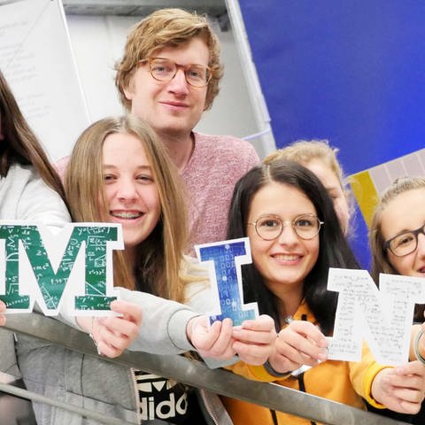 neuneinhalb-Reporter Robert zusammen mit einigen Schülerinnen im Schüler-Forschungslabor des DLR. Die Mädels halten die Buchstaben M – I – N – T in die Kamera. (Foto: WDR, tvision)