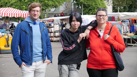 neuneinhalb-Reporter Robert steht mit dem 14-jährigen Alex und seiner Mutter Mel auf einem Wochenmarkt. Alex lehnt sich mit einem Arm auf die Schulter seiner Mutter. (Foto: WDR, tvision)