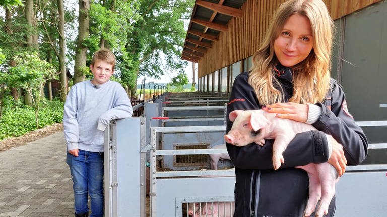 neuneinhalb-Reporterin Jana und der 9-jährige Hinnerk stehen vor den Schweineställen. Jana hält ein kleines Ferkel im Arm. (Foto: WDR, tvision)