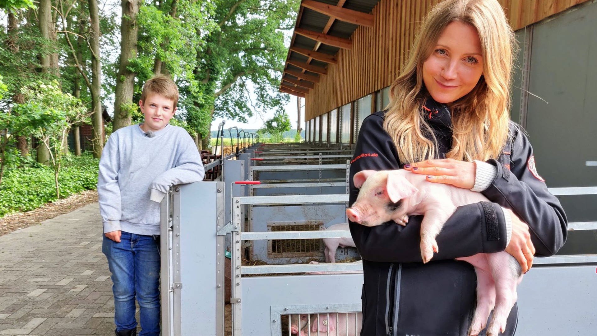 neuneinhalb-Reporterin Jana und der 9-jährige Hinnerk stehen vor den Schweineställen. Jana hält ein kleines Ferkel im Arm. (Foto: WDR, tvision)