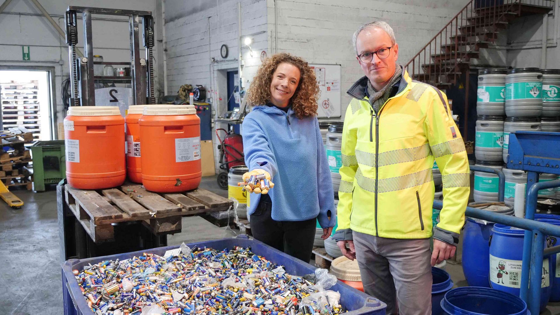 neuneinhalb-Reporterin Gesa und Florian Clever stehen vor einer großen blauen Plastikwanne voller Altbatterien in einer Recycling-Anlage. Im Hintergrund stehen bunte Plastikfässer voller Batterien. Gesa hält eine Handvoll Batterien in Richtung Kamera.
