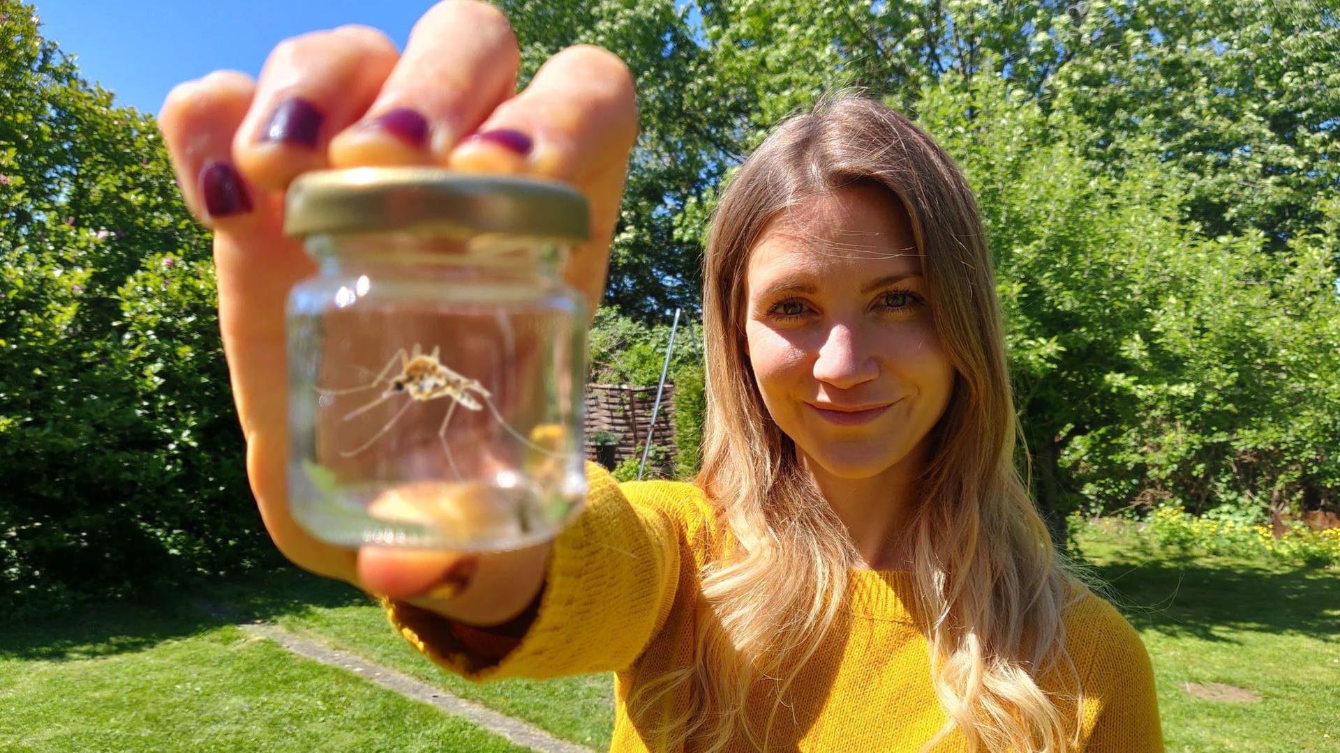 neuneinhalb-Reporterin Jana steht im Garten und zeigt eine Mücke in einem Glas