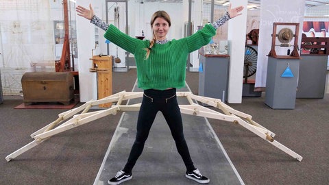 neuneinhalb-Reporterin Jana in der da Vinci Manufaktur der FH Bielefeld. Sie steht wie der vitruvianische Mensch vor einer hölzernen Leonardo-Brücke, die ganz ohne Nägel oder Schnüre gebaut wird.