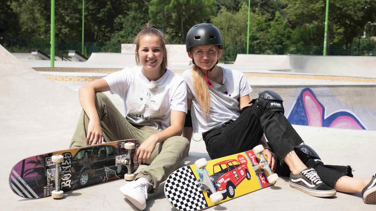 neuneinhalb-Reporterin Jana und Lea sitzen mit ihren Skateboards auf einer Skateboard-Rampe. 
