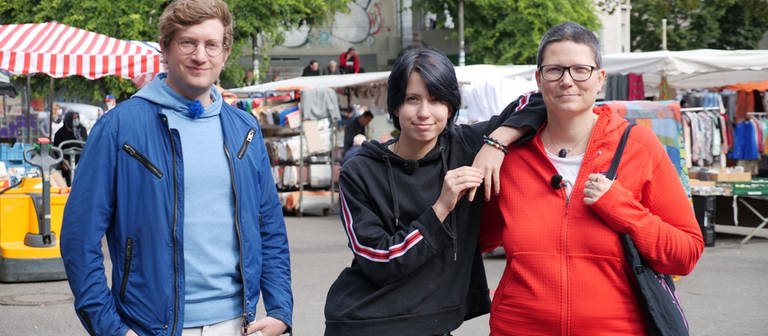 neuneinhalb-Reporter Robert steht mit dem 14-jährigen Alex und seiner Mutter Mel auf einem Wochenmarkt. Alex lehnt sich mit einem Arm auf die Schulter seiner Mutter. (Foto: WDR, tvision)