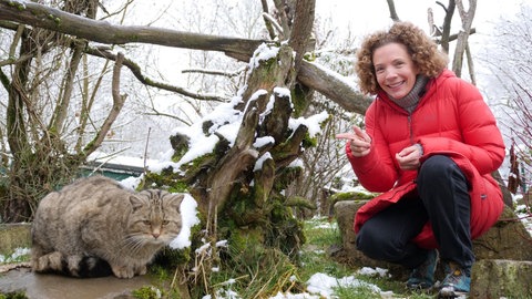 neuneinhalb-Reporterin Gesa hockt in einem Gehege im Wildkatzendorf im thüringischen Hütscheroda. Sie zeigt auf eine Wildkatze, die neben ihr sitzt.