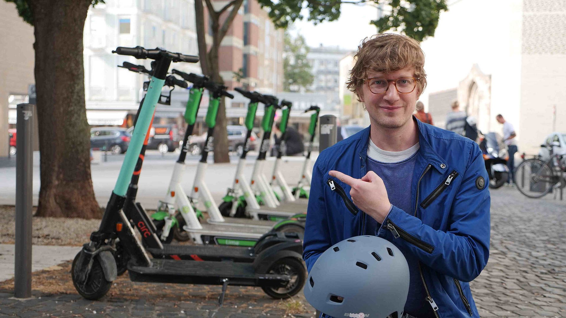 neuneinhalb-Reporter Robert steht in einer Fußgängerzone und trägt einen Fahrradhelm in der Hand. Mit der anderen zeigt er auf eine Reihe Elektro-Roller, die hinter ihm stehen.