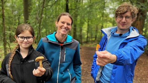 neuneinhalb-Reporter Robert mit Johanna und ihrer Mutter Melanie im Wald. Johanna und Robert halten Pilze in die Kamera.