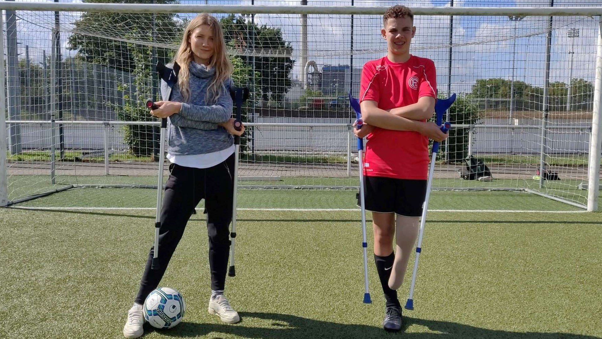 neuneinhalb-Reporterin Jana und der 14-Jährige Jamie stehen mit Krücken in den Händen vor einem Fußballtor. Jamie ist Amputierten-Fußballer, ihm fehlt ein Bein.