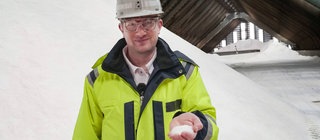 Robert steht in einer Halle, in der Salz aufgetürmt ist. Er trägt Schutzkleidung und hält Salz in der Hand.