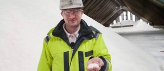 Robert steht in einer Halle, in der Salz aufgetürmt ist. Er trägt Schutzkleidung und hält Salz in der Hand.