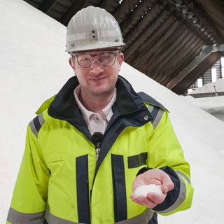 Robert steht in einer Halle, in der Salz aufgetürmt ist. Er trägt Schutzkleidung und hält Salz in der Hand.