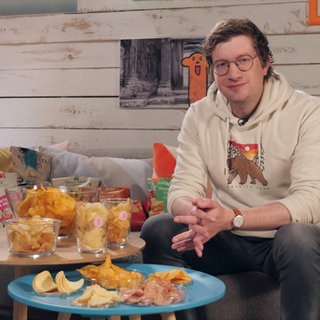 Robert sitzt im neuneinhalb-Studio. Vor ihm stehen zwei Tische mit Schalen voller Chips.