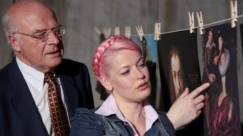 Enie van der Meiklokjes und Prof. Raimund Wünsche stehen vor einer Wäscheleine, an der Gemälde aufgehängt sind.