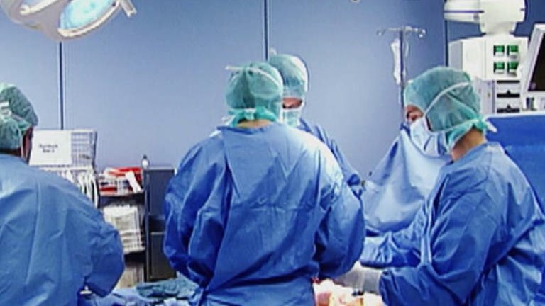 Mehrere Ärzte in blauen Kitteln stehen um einen Patienten. (Foto: SWR – Screenshot aus der Sendung)