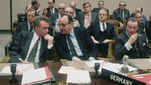 Sitzungssaal: Verteidigungsminister Hans Apel (links) am Tisch neben Außenminister Hans-Dietrich Genscher (rechts); dahinter Vertreter anderer Nationen (Foto: SWR - Screenshot aus der Sendung)
