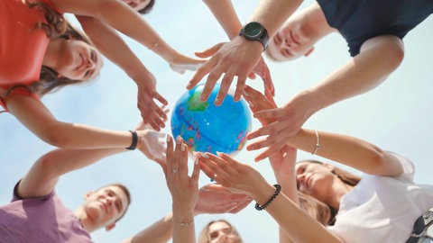 Symbolbild für Sport und Spiele auf der ganzen Welt: Mehrere Personen stehen im Kreis strecken die Arme nach oben und halten gemeinsam einen Globus. Das Bild ist von unten fotografiert.