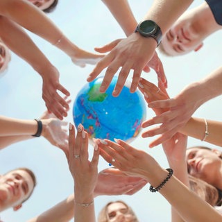 Symbolbild für Sport und Spiele auf der ganzen Welt: Mehrere Personen stehen im Kreis strecken die Arme nach oben und halten gemeinsam einen Globus. Das Bild ist von unten fotografiert. 