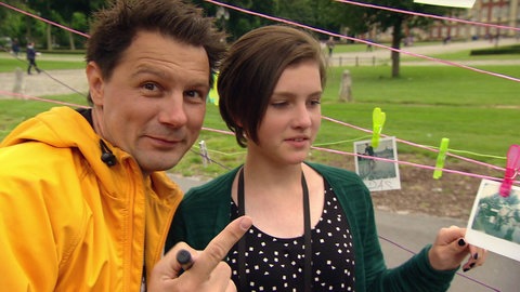 Moderator Andre Gatzke neben dem Mädchen Lily, hinter ihnen hängen Fotos an einer Wäscheleine.