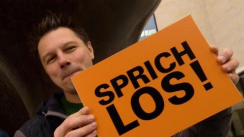 Moderator André Gatzke mit Deutschlernenden, die Schilder in der Hand halten mit der Aufschrift 