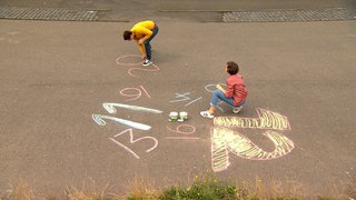 Ein Mädchen und ein junger Mann malen mit Kreide Zahlen auf den Boden.