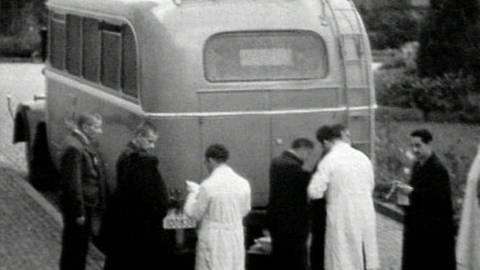 Ein schwarz-weiß Foto eines Buses, vor dem mehrere Menschen in Anzügen und Laborkitteln stehen.