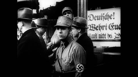 Mann in Uniform mit Hakenkreuz-Armbinde vor Schild "Deutsche wehrt euch. Kauft nicht bei Juden”
