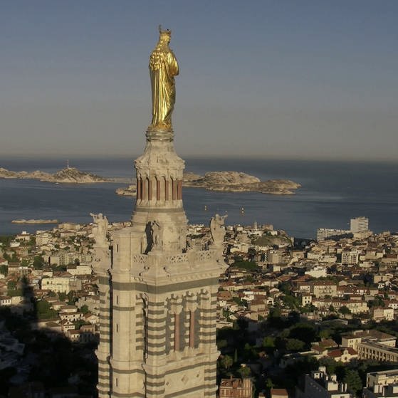 Luftaufnahme von Marseille mit einer goldenen Skulptur im Vordergrund, die hoch über der Stadt auf einem Turm steht.
