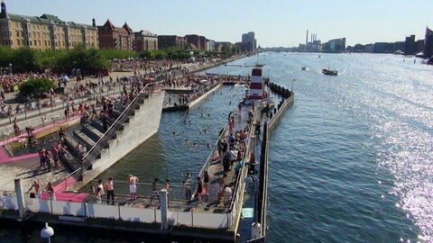 Man sieht ein Schwimmbad, das direkt neben einen Fluss gebaut und voller Mensch ist.