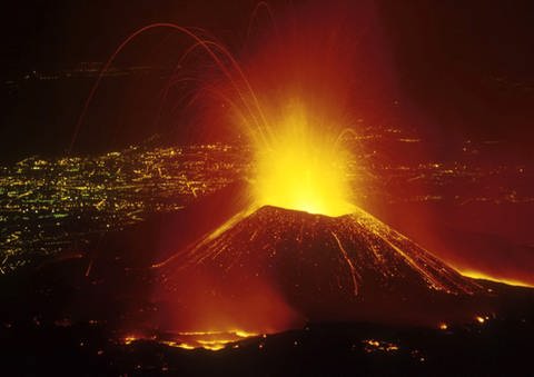 Nächtlicher Blick von seitlich oben auf den Vulkan Ätna, der gerade Feuer speit. Im Hintergrund die Lichter der Großstadt Catania. (Foto: AKG)