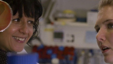 Eine Forscherin hält eine Petrischale in die Höhe und zeigt sie einer Frau rechts.