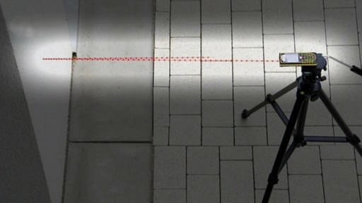 Ein Laserscanner steht auf einem Kachelfußboden, ein roter Strahl trifft auf die Wand. (Foto: SWR – Screenshot aus der Sendung)