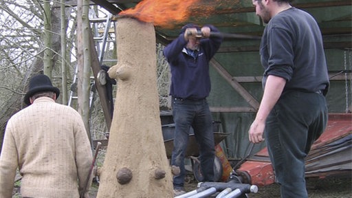 Drei Männer stehen um einen mannshohen Rennofen aus Lehm. Am oberen Ende schlägt Feuer aus der Konstruktion.