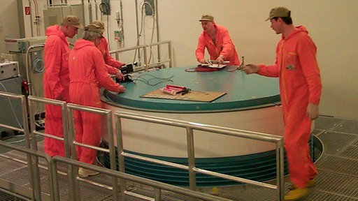 Fünf Männer in roten Overalls stehen um einen großen runden Behälter.