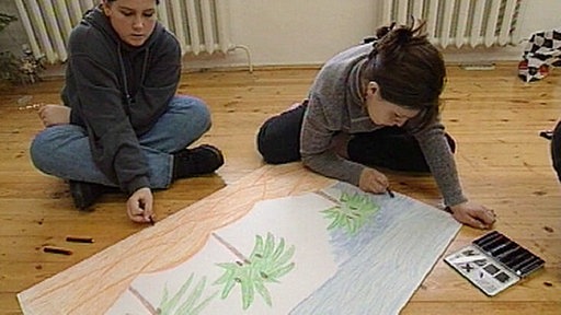 Zwei Kinder sitzen auf dem Boden und zeichnen an einem großen Bild, auf dem Palmen und Strand zu sehen sind.