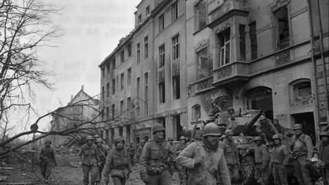Soldaten in Uniform laufen an einer ausgebrannten Häuserfront entlang, vor den Häusern steht ein Panzer (Foto: National Archive)