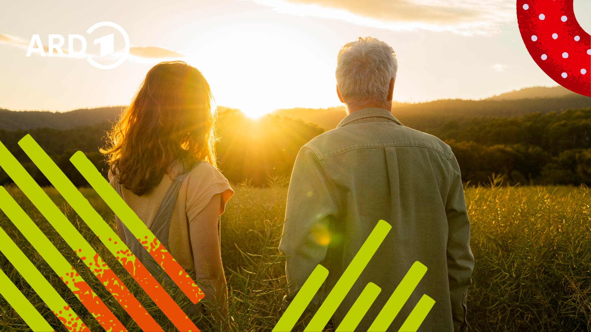 Ein Vater steht mit seiner Tochter in einem Feld und sieht einem Sonnenuntergang zu.