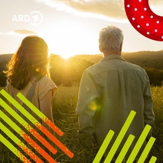 Ein Vater steht mit seiner Tochter in einem Feld und sieht einem Sonnenuntergang zu.