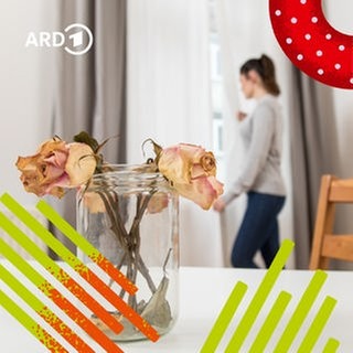 Zwei verwelkte Rosen in einer Vase auf einem Esstisch und eine Frau im Hintergrund, die traurig aus dem Fenster sieht.