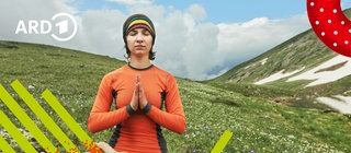 Eine Frau sitzt im Schneidersitz mit geschlossenen Augen und gefalteten Händen vor der Brust auf einem Berg und meditiert.