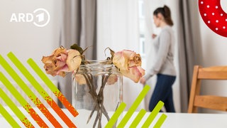 Zwei verwelkte Rosen in einer Vase auf einem Esstisch und eine Frau im Hintergrund, die traurig aus dem Fenster sieht.