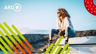 Eine Frau sitzt auf einem Camper am Meer und sieht glücklich in die Ferne.